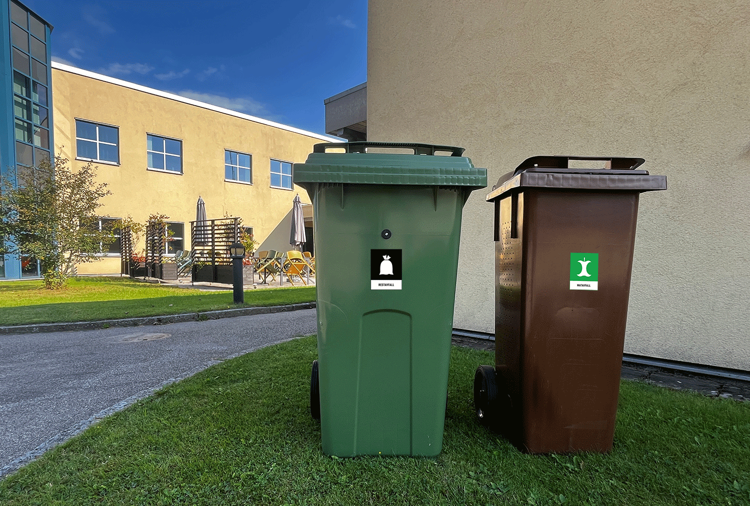 Gron_brun_karl Ett grönt och ett brunt avfallskärl står på en gräsmatta framför en kontorsbyggnad