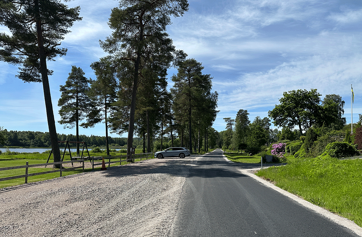 En nyasfalterad väg i Mogdens Fritidshusområde. Sjö, tallar och gräs vid sidan.